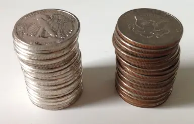 Two stacks of half dollars side by side on a white surface. The edges of the stack on the left are solid silver-white; every edge in the stack on the right shows a brown copper stripe.